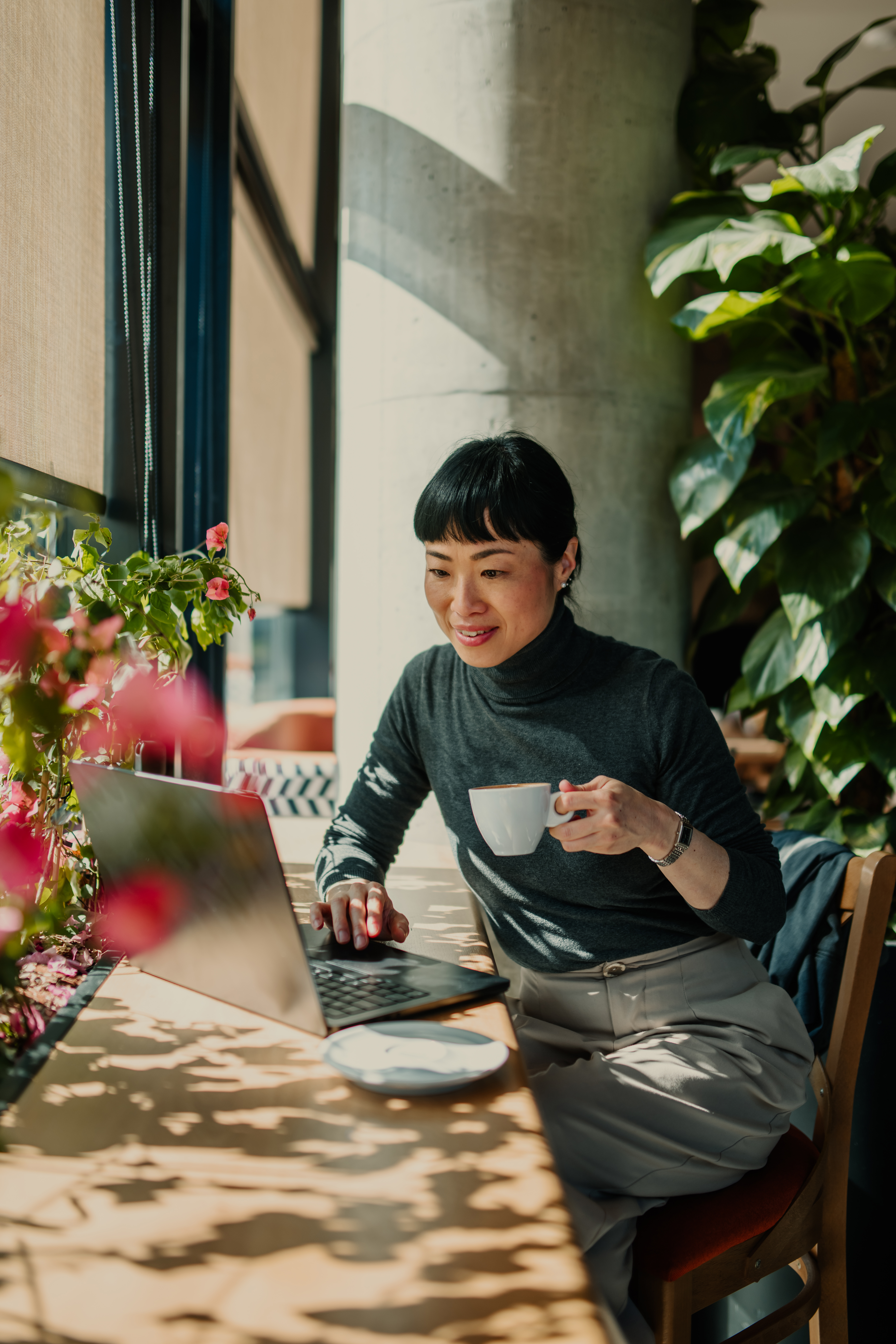 Asian businesswoman enjoying a coffee while working on her laptop in a bright and sunny cafe